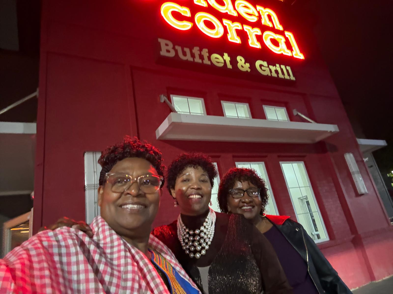 Three community members smiling beneath a restaurant sign at night